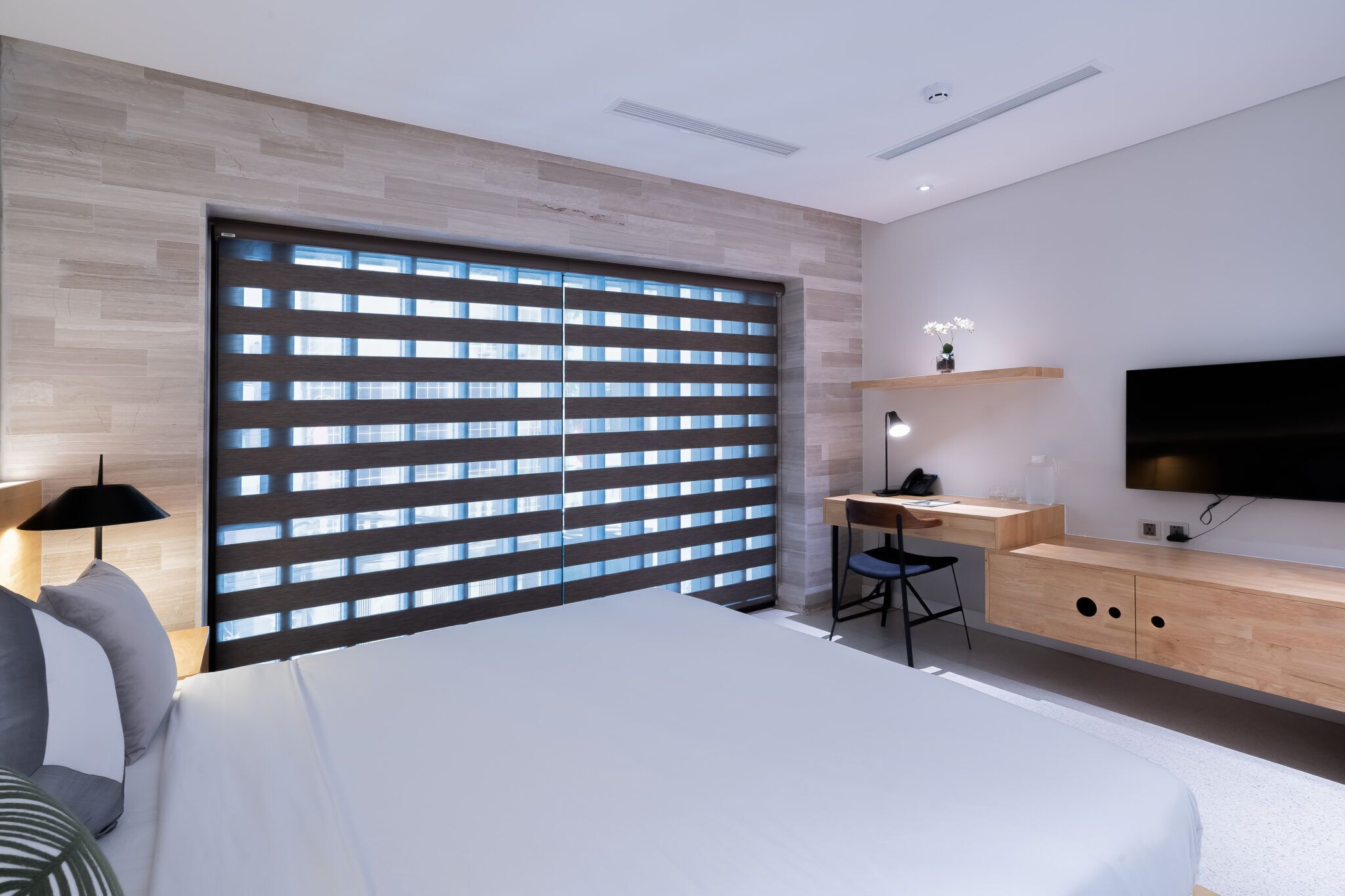A modern bedroom with a large bed, a desk, a chair, a TV, and a window with blinds.