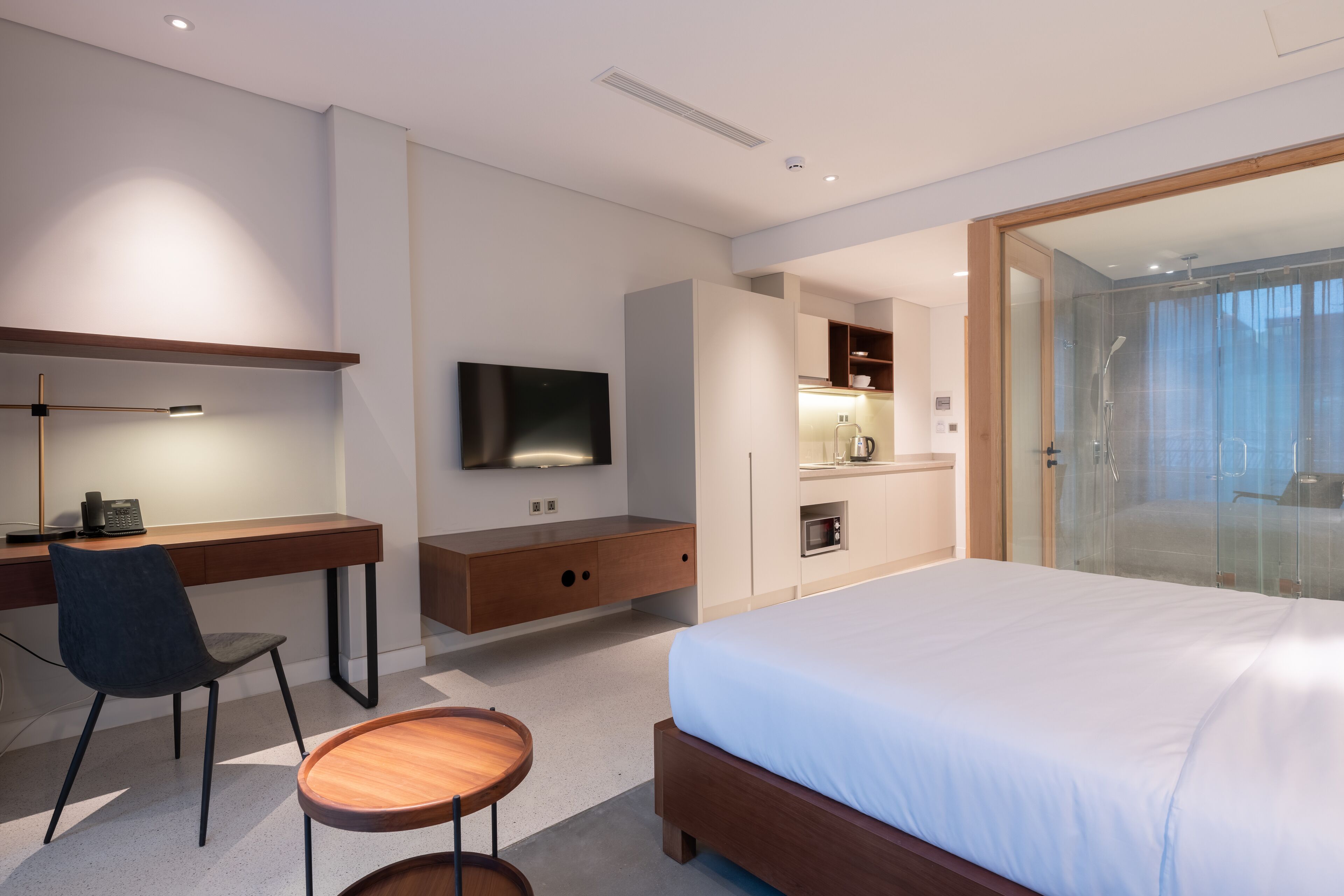 A modern hotel room with a large bed, a desk with a chair, a television, a small kitchen area, and a glass-enclosed shower.