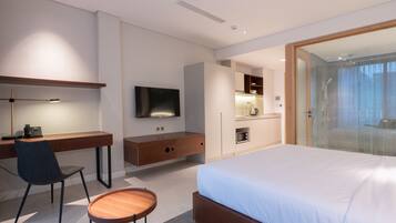 A modern hotel room with a large bed, a desk with a chair, a television, a small kitchen area, and a glass-enclosed shower.