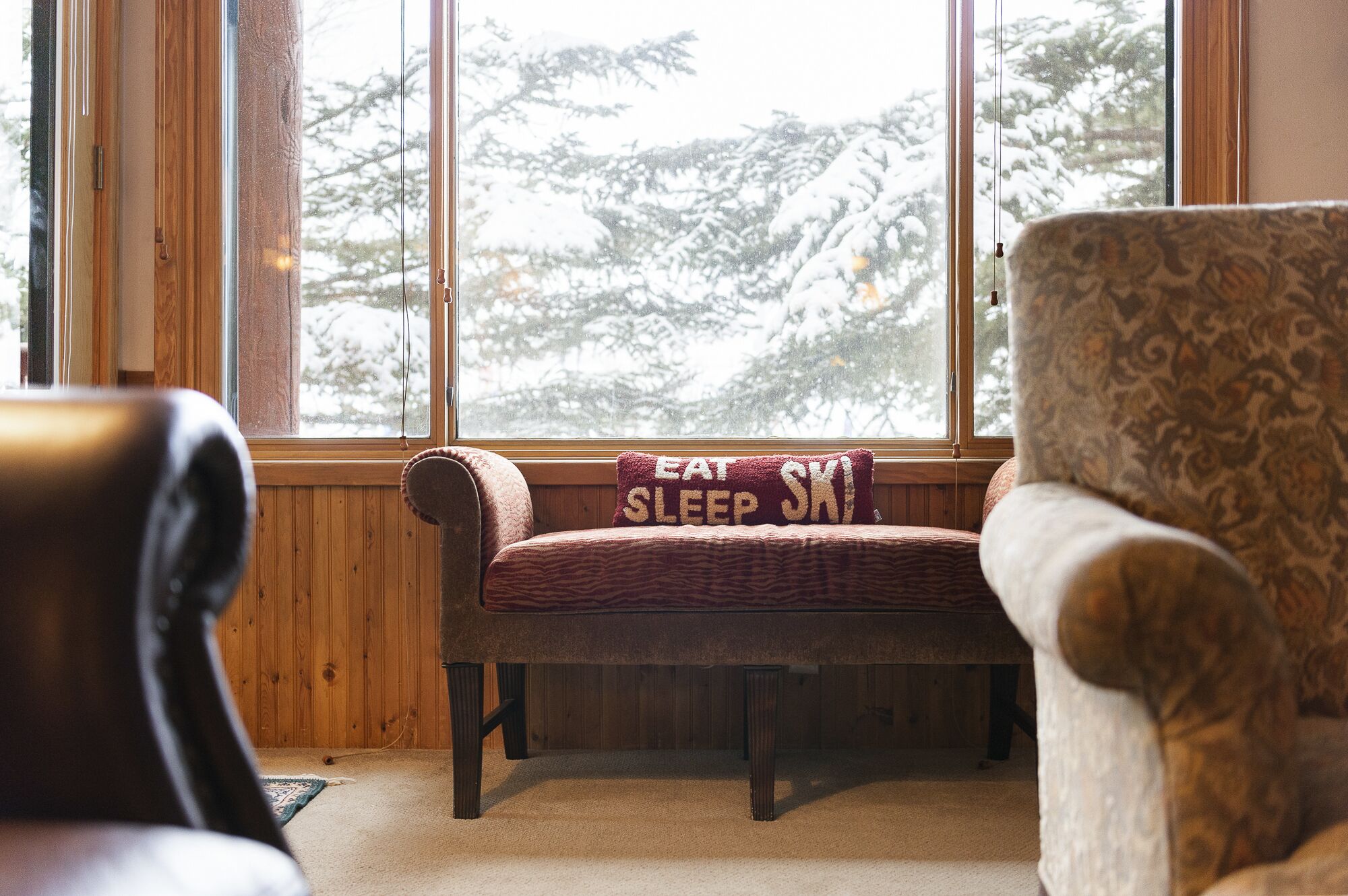 Een gezellige kamer met een houten bankje en een stoel bij een raam met uitzicht op besneeuwde bomen.
