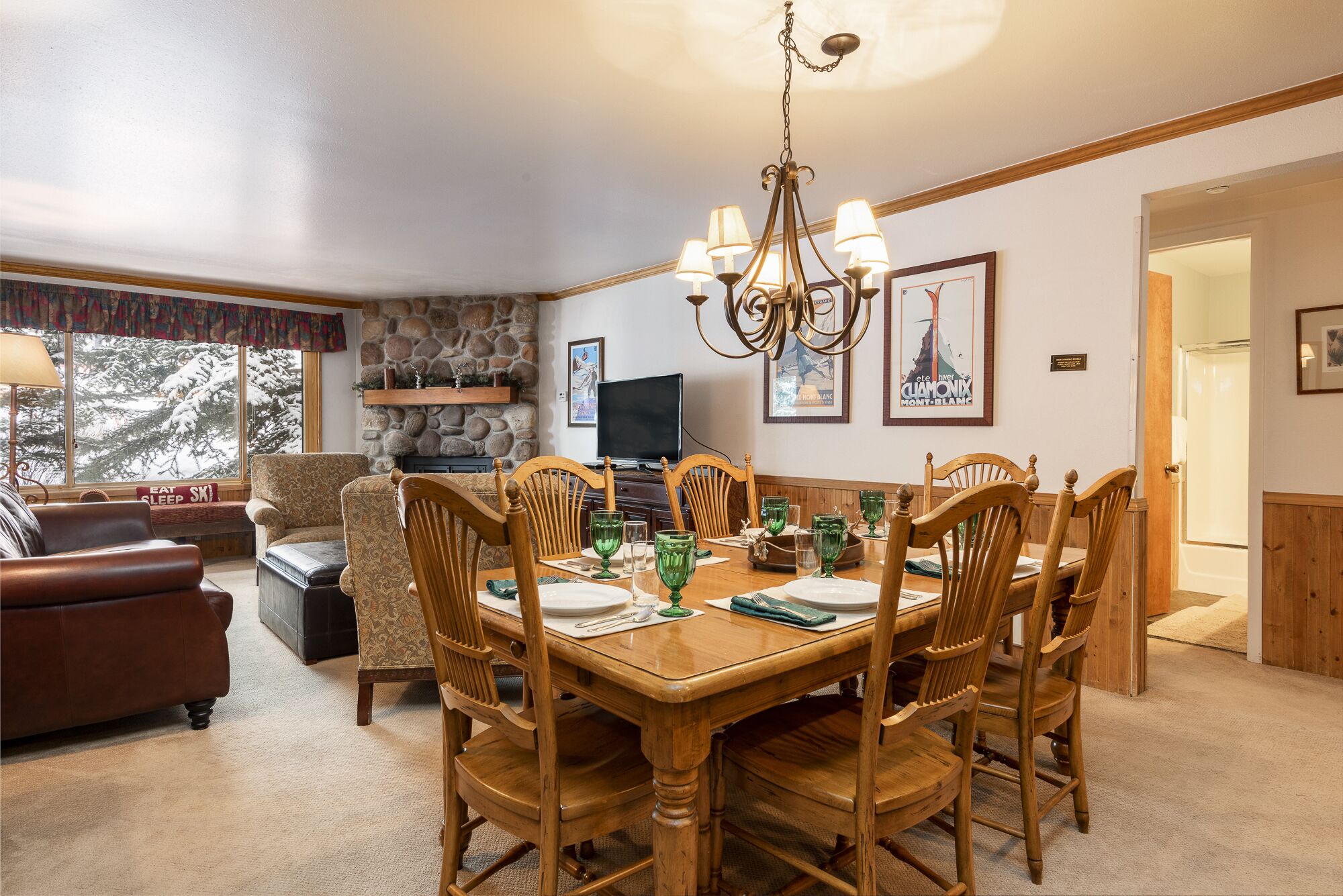 A dining room with a wooden table set for six, surrounded by six wooden chairs. A stone fireplace with a television on top is visible in the background.