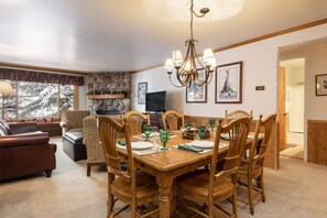 A dining room with a wooden table set for six, surrounded by six wooden chairs. A stone fireplace with a television on top is visible in the background.