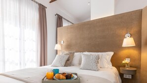 En pænt redt seng med et bakke med morgenmad, en sengetavlelampe og udsigt til byen gennem tynde gardiner.