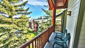 A balcony with wooden railings overlooking a lush green landscape and a building with a red balcony. - Managed by Park City Lodging-True Ski-In/Ski-Out to Park City Mountain Resort! (Park City)