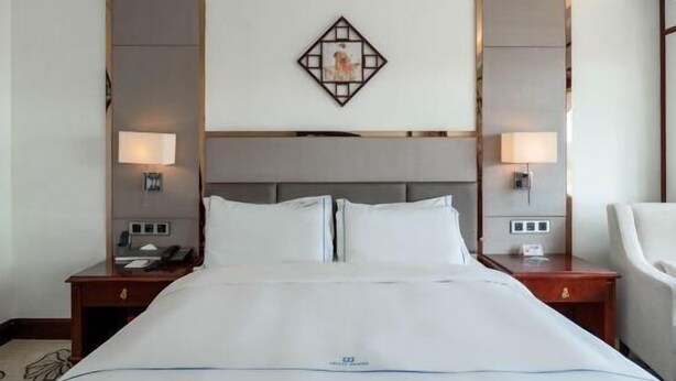 A hotel room with a large bed, two bedside tables, a nightstand, a chair, and a decorative wall piece. - Junyi Hotel (Shenzhen)