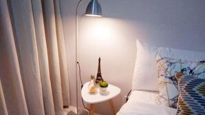 Une chambre d’hôtel bien rangée, avec un lit, une lampe de chevet, une petite table agrémentée d’objets décoratifs et un rideau.