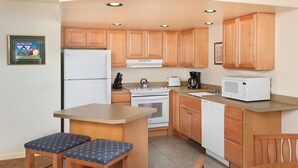 Cocina compacta con armarios de madera, un refrigerador blanco, estufa, microondas y un lavamanos. Hay dos taburetes y una mesa pequeña.