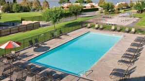 Una piscina rectangular con superficie de agua azul cristalina, rodeada por una plataforma de hormigón. Hay varias tumbonas y sombrillas a lo largo de la piscina. En el fondo, se observan colinas y una masa de agua, probablemente un lago o un río.