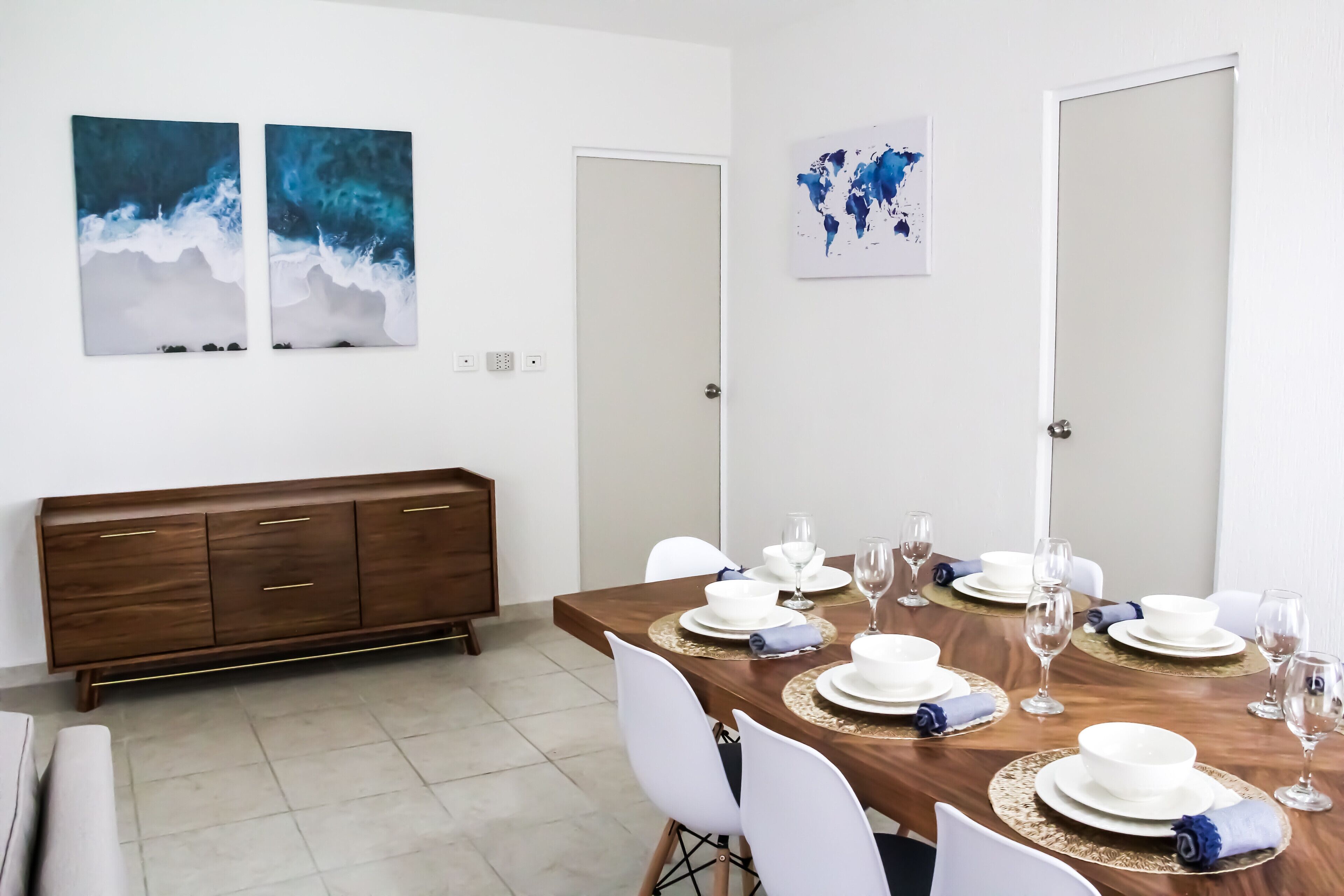 A dining area with a wooden table set for six, white chairs, and a world map on the wall.