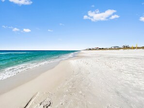 Une plage avec une eau d’un bleu limpide, un rivage sablonneux et des maisons en arrière-plan.