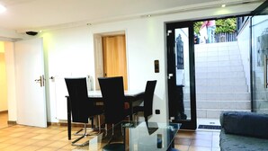 Un espace repas moderne avec une table en verre et des chaises noires, une porte en bois et une porte-fenêtre coulissante donnant sur un balcon.