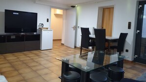 Un salon moderne avec une table à manger en verre, des chaises noires, un réfrigérateur blanc, un téléviseur à écran plat et une porte en bois.