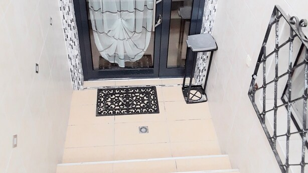 Un escalier menant à une porte surmontée d’un paillasson à motifs, une grille en métal et un mur recouvert de carreaux avec une bordure en mosaïque.