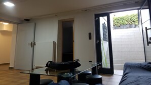 Une pièce avec une table en verre, des tabourets noirs, un radiateur et une porte donnant sur un espace extérieur agrémenté d’une grille.