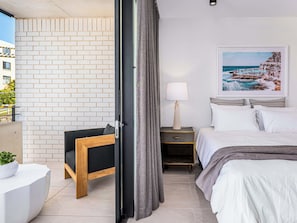 A modern hotel room with a bed, bedside table, lamp, and a view of a beach scene.