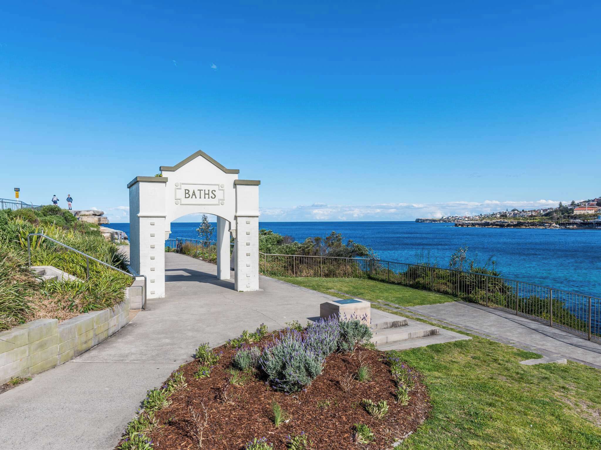 Un arc blanc portant l’inscription « BAINS », d’où l’on a une vue dégagée sur la mer et sur une ville située au loin.