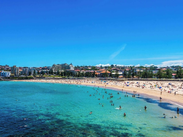 A beach with clear turquoise water, a sandy shore, and buildings in the background.