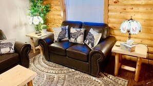 A cozy log cabin interior with a leather sofa, wooden side tables, a patterned rug, and a potted plant.