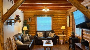 A log cabin interior with a wooden ceiling, a fireplace, a sofa, a table, and a window.