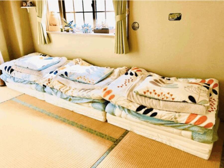 A room with a futon bed, a small window with curtains, and a wall-mounted shelf with decorative items.