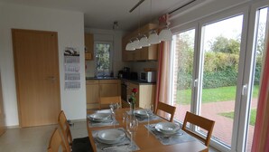 Zona pranzo con tavolo in legno apparecchiato per quattro, cucina sullo sfondo e vista giardino attraverso ampie finestre.