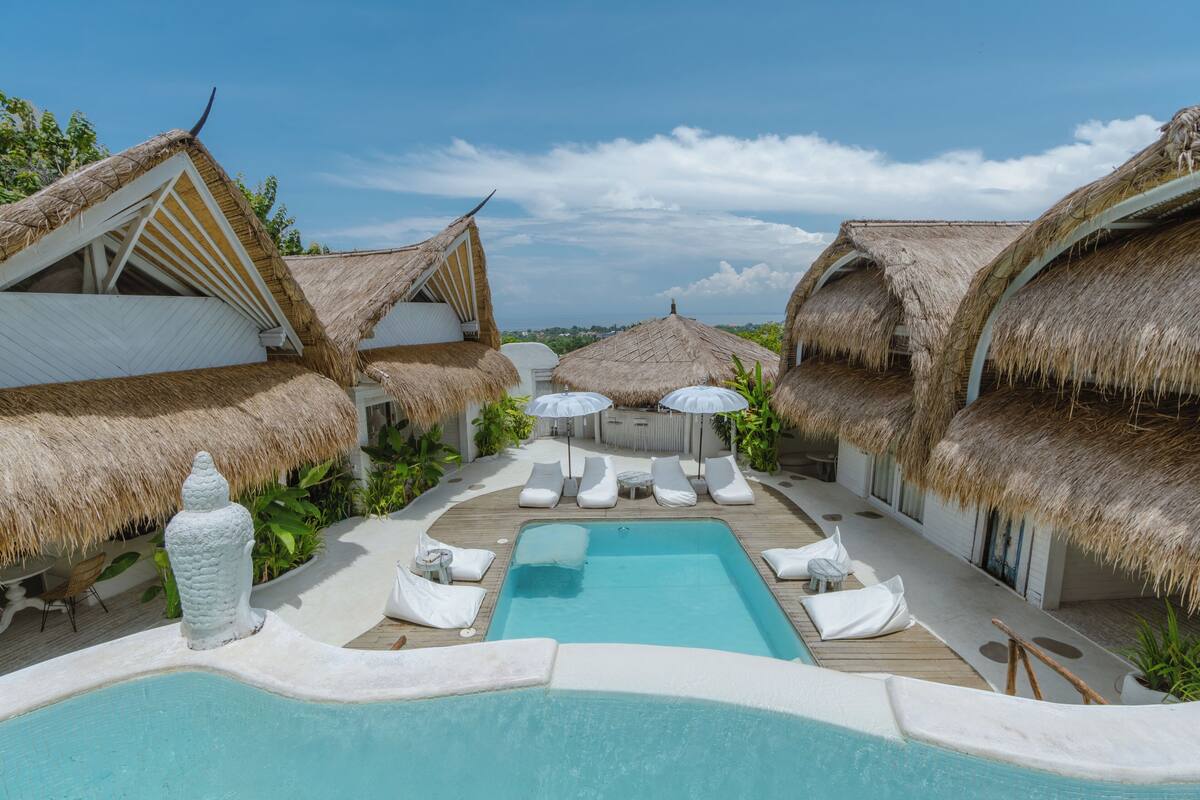 Een zwembadgedeelte met bungalows op palen onder rieten daken, ligstoelen en parasols.