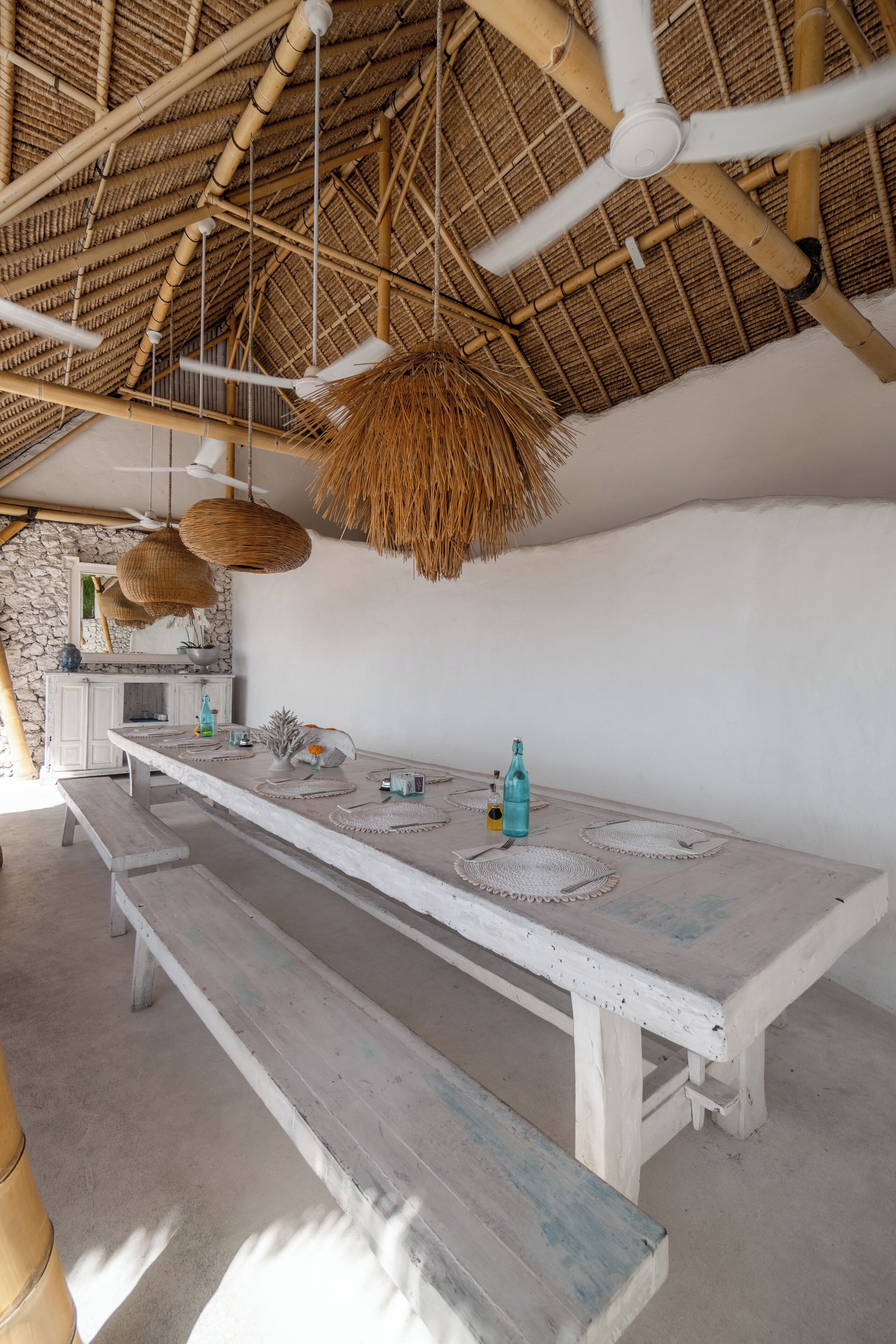A long wooden table with woven placemats and a bench, a ceiling fan, and a thatched roof.