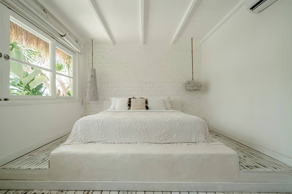 Een slaapkamer met witte bakstenen muren, een bed, een hanglamp en uitzicht op groen buiten het raam.