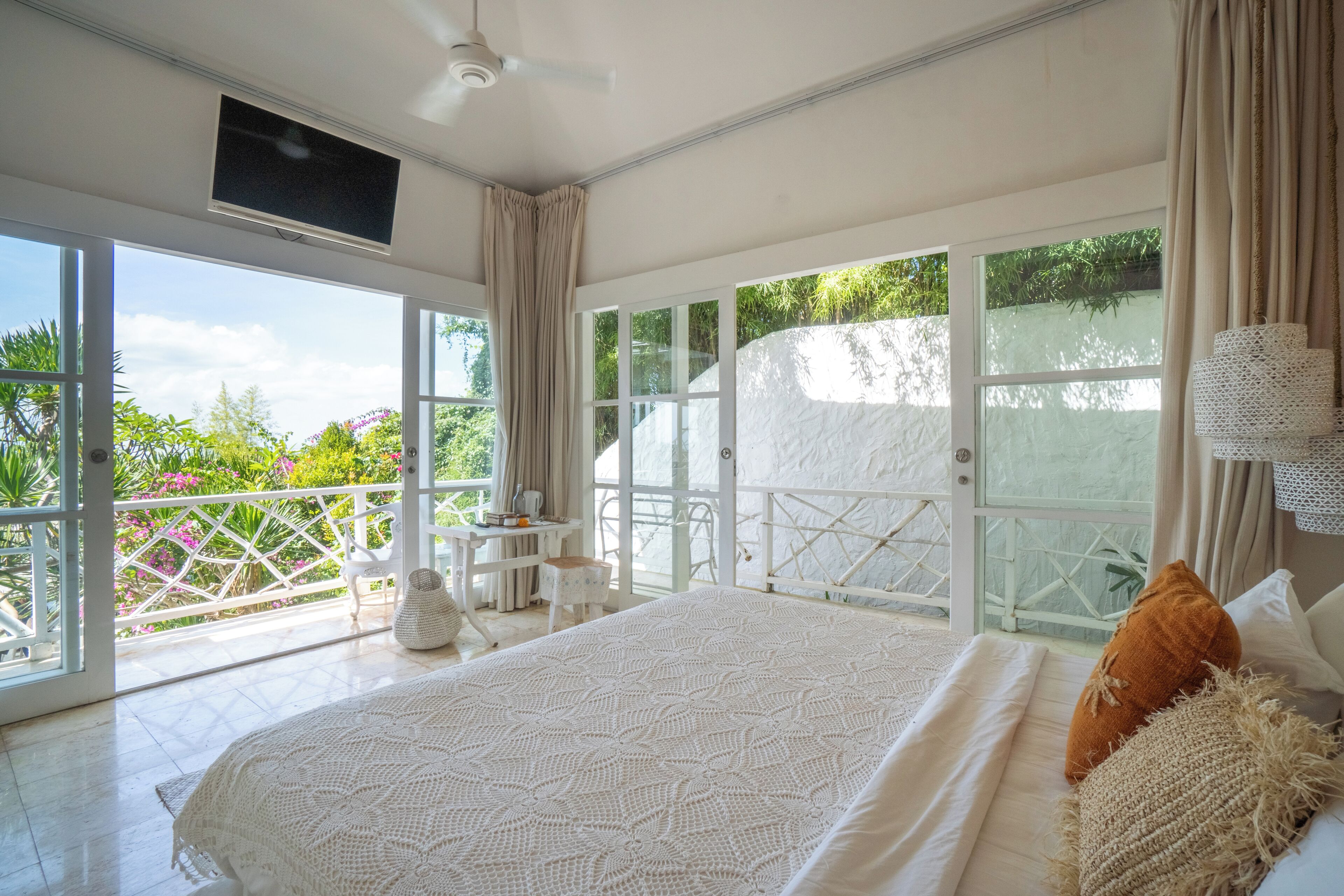 A spacious bedroom with a large bed, a ceiling fan, and a view of a lush garden through large sliding glass doors.