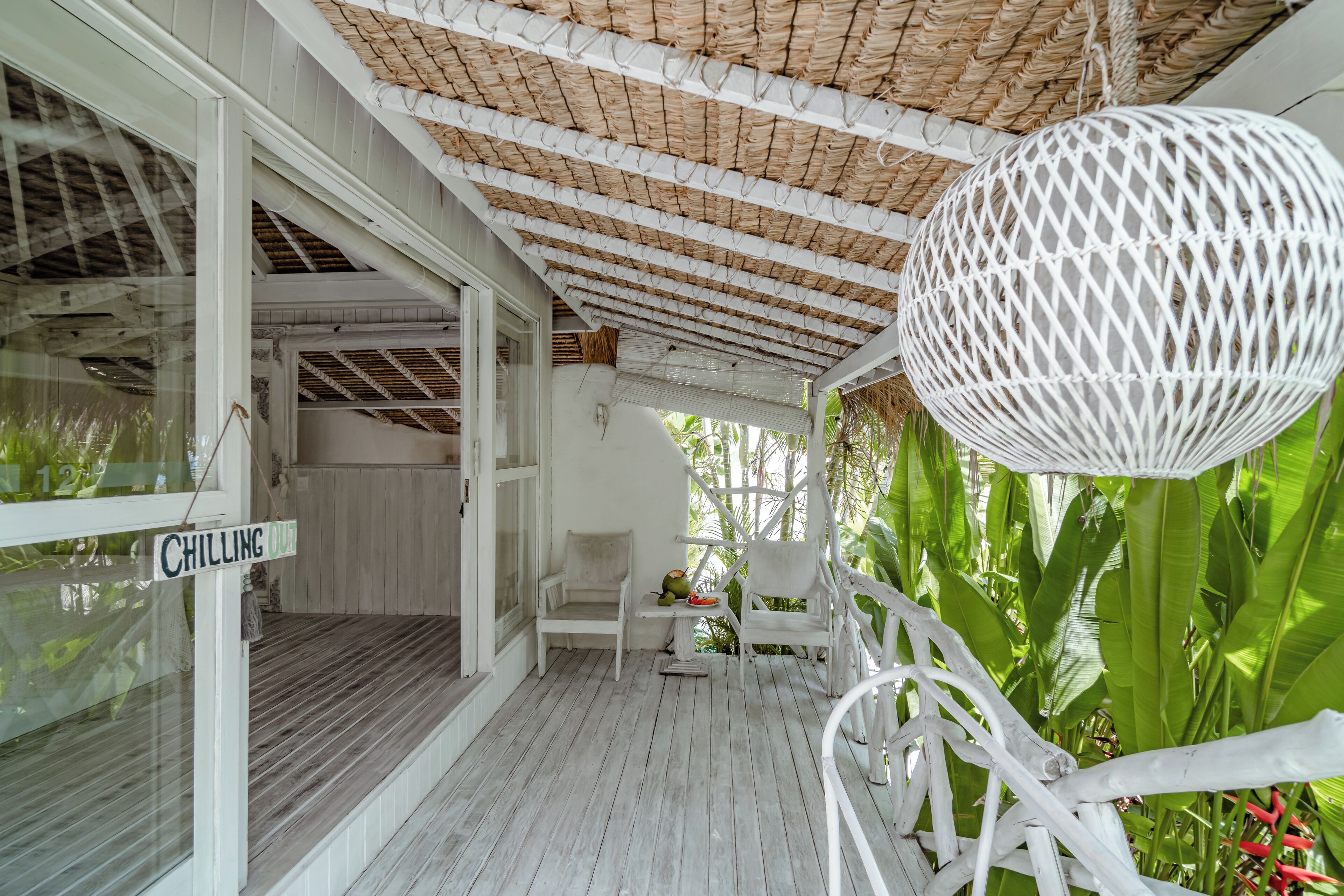 A porch with white wicker furniture, a hanging white lantern, and lush green foliage.