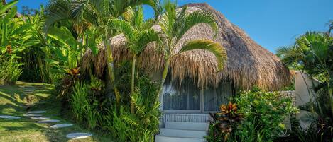 A thatched-roof building with a stone staircase leading up to a porch, surrounded by lush tropical vegetation.