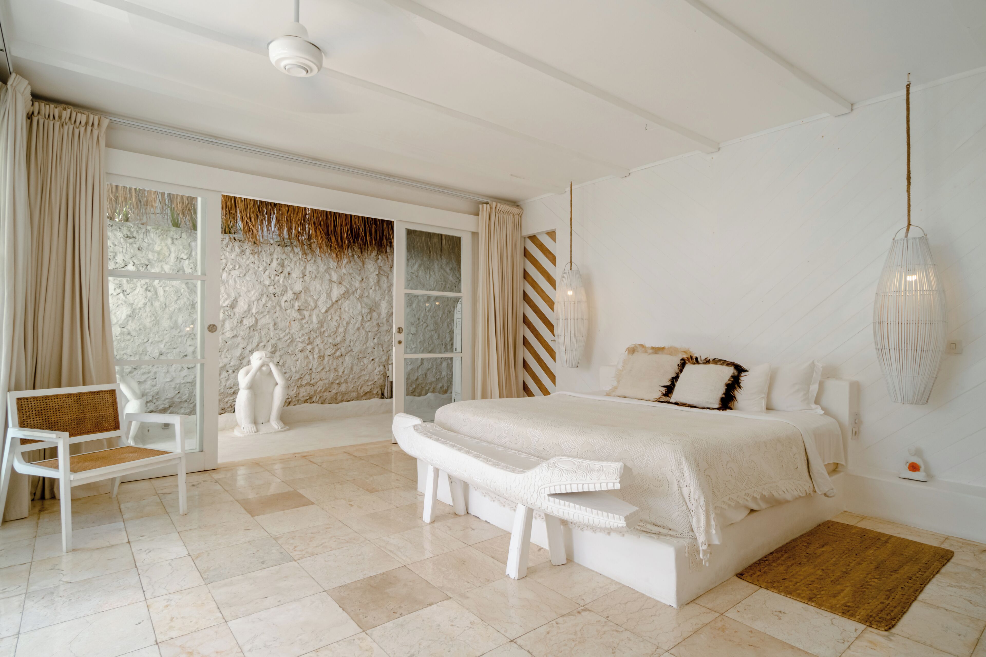 Un dormitorio con una cama grande, un banco, una silla y vistas a través de un ventanal.