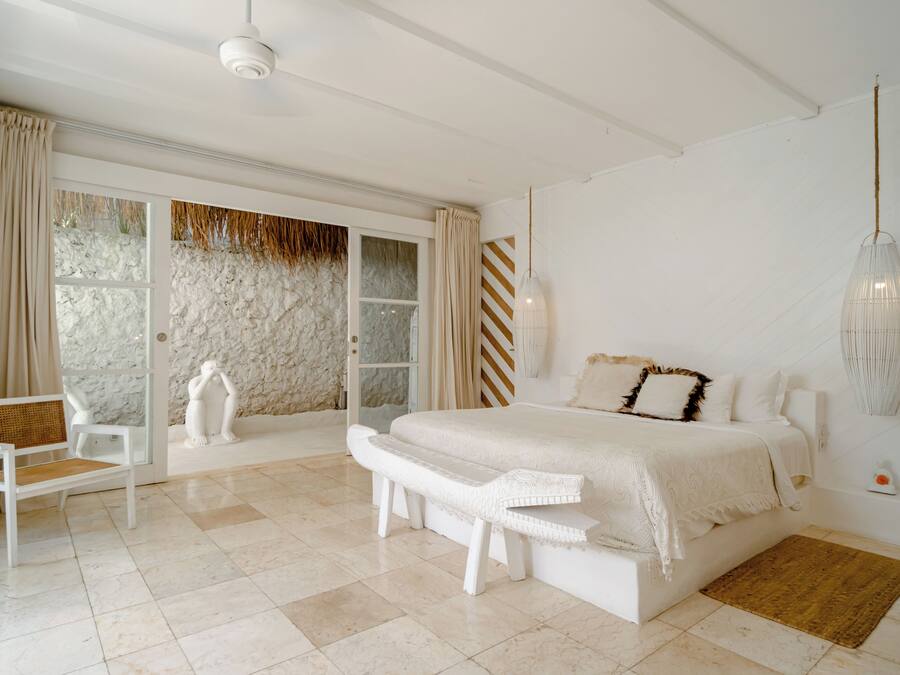 Un dormitorio con una cama grande, un banco, una silla y vistas a través de un ventanal.