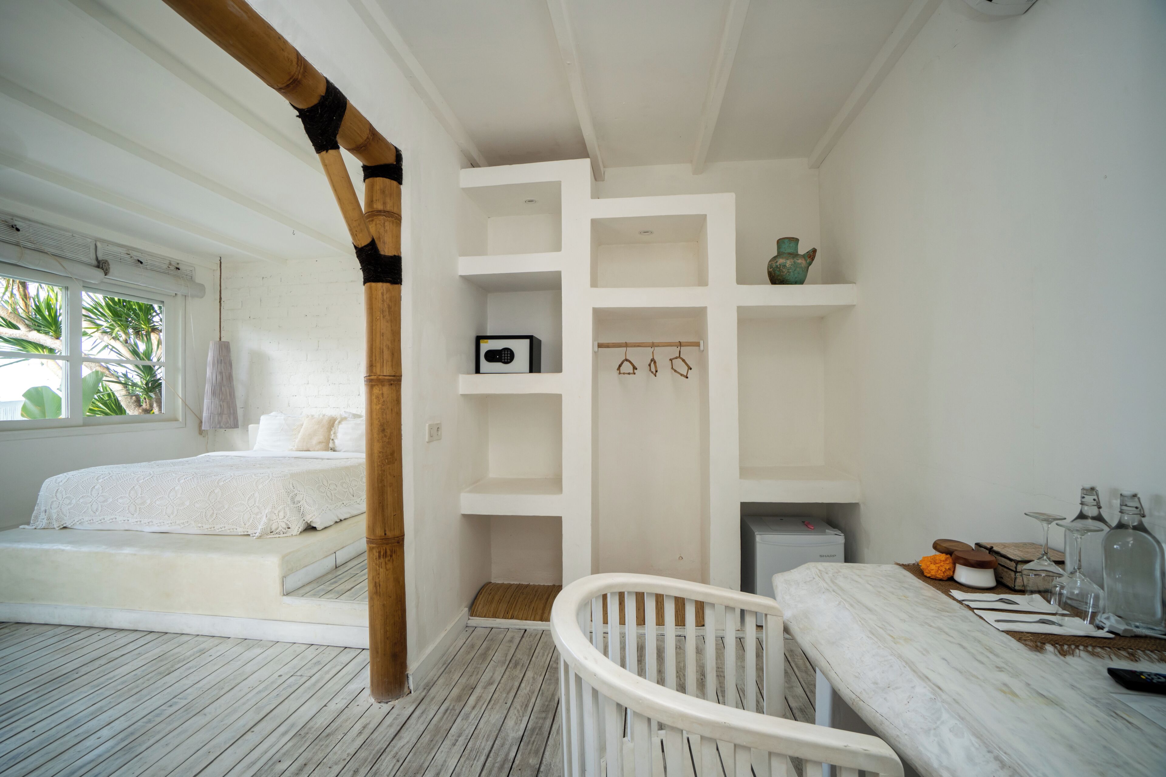 A bedroom with a bed, a white crib, a shelf with decorative items, and a window with a view of palm trees.