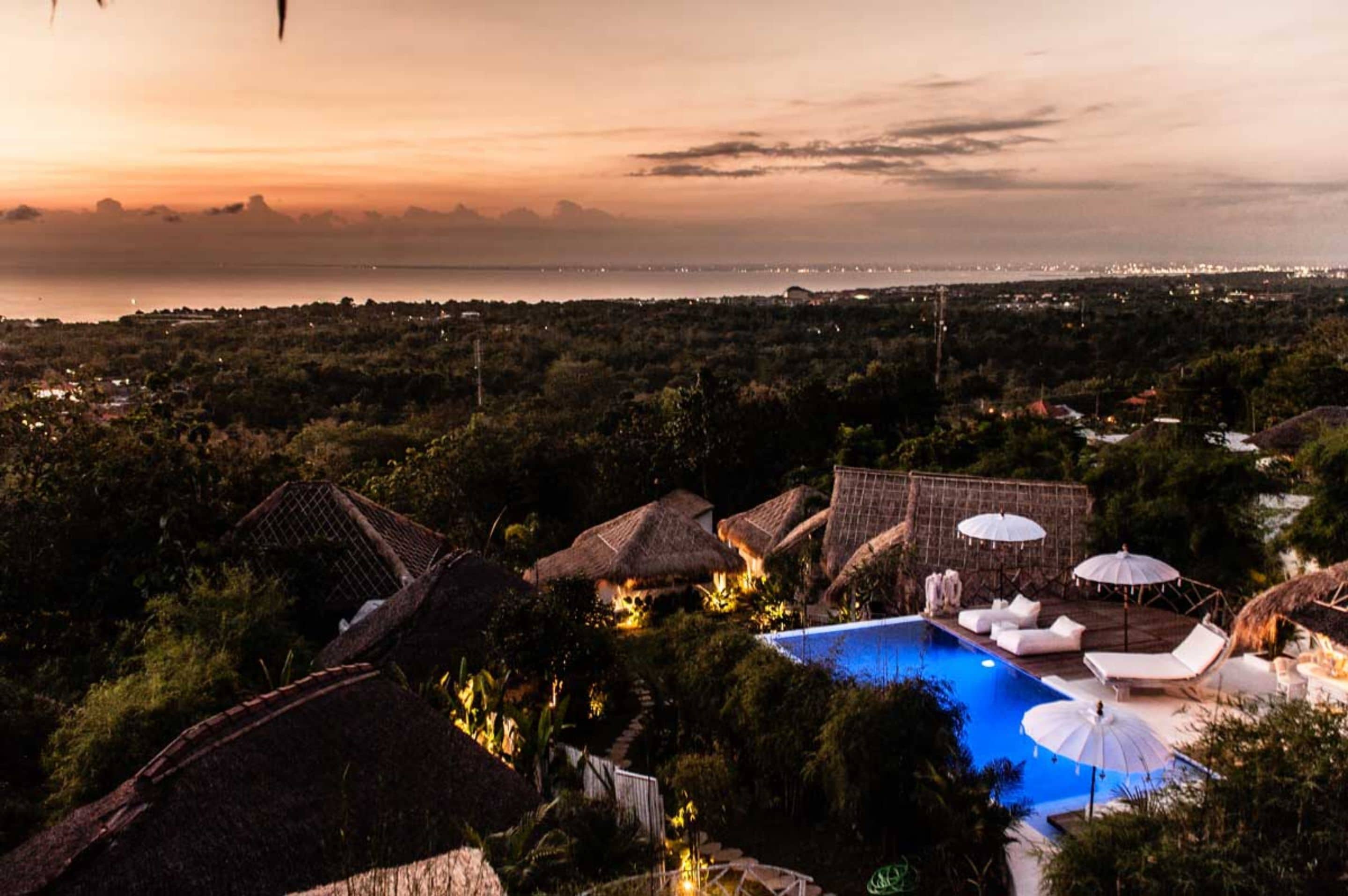 Un resort con piscina, construcciones de techo de paja y vistas al océano al atardecer.
