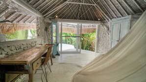 Una habitación con un escritorio de madera, una silla, una cama con sábana blanca, una pared de piedra, un techo de paja y vistas a la vegetación exterior.