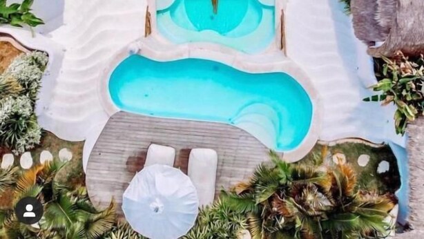 Un área de piscina con una piscina rectangular central, rodeada de tumbonas y sombrillas, y plantas tropicales.