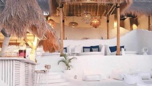 Un área de descanso frente a la playa con estructuras de techo de paja, sillas de lounge blancas y una zona de bar.