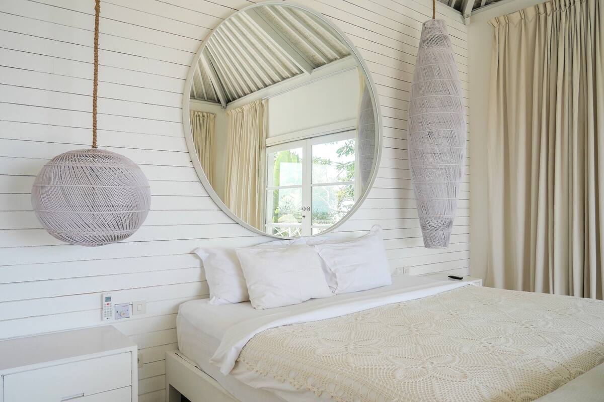 Een slaapkamer met een bed, een ronde spiegel, een hanglamp en een raam met gordijnen.