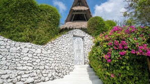 Un sendero de piedra conduce a una estructura tradicional con techo de paja, rodeada de exuberante follaje y vibrantes flores rosas.