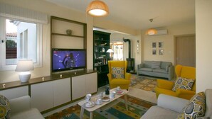 Ein Wohnzimmer mit Fernseher, gelben Ohrensesseln, einem grauen Sofa und einem Couchtisch mit Tassen und Untertassen.