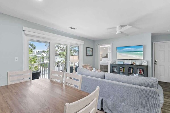 A dining area with a wooden table and chairs, a grey-striped sofa, a television, and a bookshelf.