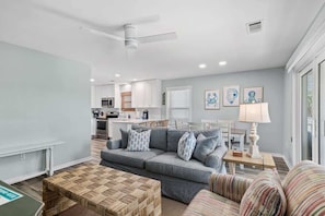 A living room with a gray sofa, a woven coffee table, and a striped armchair. A kitchen is visible in the background with white cabinets and a dining area.