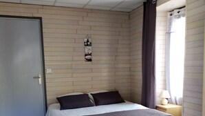 Un dormitorio con cama, mesita de noche, lámpara de pared y ventana con cortinas.