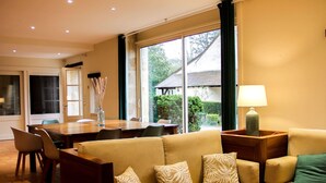 Un salon avec un canapé beige, une table à manger et une vue sur un jardin, visible à travers de grandes fenêtres.