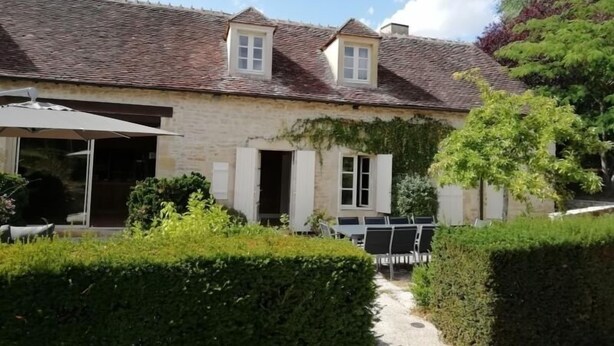 Une maison en pierre avec un toit en tuiles, un patio couvert et un jardin agrémenté de haies.