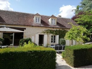 A stone house with a tiled roof, a covered patio, and a garden with hedges.