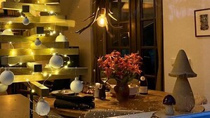Ein gemütliches Zimmer mit einem mit Lichtern und Verzierungen geschmückten Holz-Weihnachtsbaum, einer Hirsch-Dekoration und einem Tisch mit Blumen in einer Vase und einer Lampe.