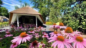 Ein strohgedecktes Pavillon, umgeben von leuchtend violetten und rosa Blumen, mit einem üppigen Garten und Bäumen im Hintergrund.