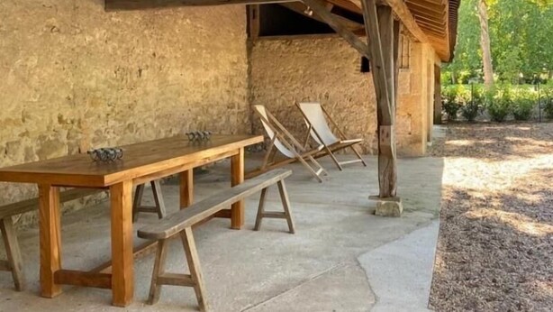 A rustic outdoor seating area with wooden benches and tables, set against a stone wall and surrounded by greenery.